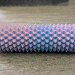 Puede incluir: Objeto cil&iacute;ndrico texturizado con un degradado de tonos rosa y azul, cubierto de p&uacute;as c&oacute;nicas espaciadas uniformemente. El objeto tiene un extremo verde y estriado. Otros objetos similares son visibles en el fondo.