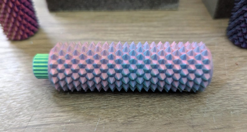 Puede incluir: Objeto cil&iacute;ndrico texturizado con un degradado de tonos rosa y azul, cubierto de p&uacute;as c&oacute;nicas espaciadas uniformemente. El objeto tiene un extremo verde y estriado. Otros objetos similares son visibles en el fondo.