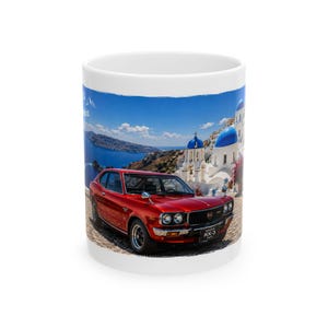May include: A white ceramic mug featuring a printed image of a red 1975 Mazda RX-3 car parked on a cobblestone path. The background displays a scenic coastal landscape with white buildings, blue domes, and a clear blue sky over the ocean.