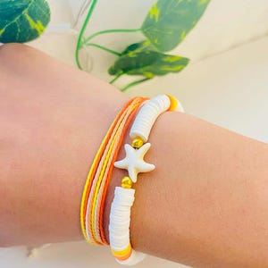 May include: A stackable bracelet set featuring a white starfish charm with gold accents. The set includes a white, orange, and yellow beaded bracelet and a multi-strand bracelet with matching colors. The bracelets are designed for a beach or summer theme.
