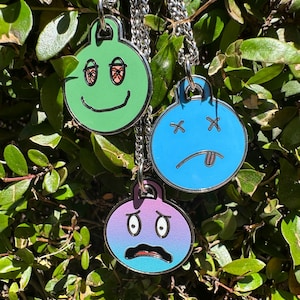Puede incluir: Tres collares con colgantes redondos y coloridos con caras de dibujos animados. Los colgantes son verdes, azules y un degradado de morado y azul. Cada colgante está unido a una cadena plateada y cuelga sobre un fondo de hojas verdes.