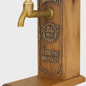 Op de afbeelding: Houten whiskydispenser met een messing kraan. De dispenser heeft het Jack Daniel's logo en de tekst "Old No. 7 Brand Tennessee Whiskey", in het hout gegraveerd. Het heeft een rechthoekige vorm met een voet.