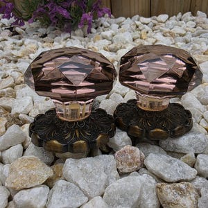 May include: A pair of antique door knobs featuring faceted, rose-tinted glass tops. The knobs are mounted on ornate, dark bronze-coloured bases. They are displayed on a bed of white and tan stones, with purple flowers in the background.