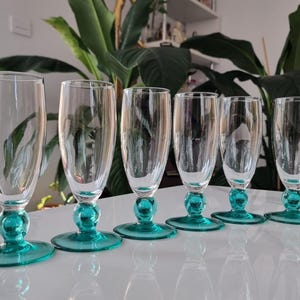 May include: A set of six clear champagne flutes. Each flute has a teal-coloured base and stem. The glasses have a classic design with a rounded base and a long, slender bowl. The teal adds a pop of colour to the glassware.