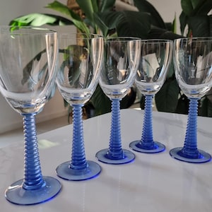 May include: Five clear wine glasses with blue, spiral-textured stems and round bases. The glasses have a classic tulip shape. The background features green plants. The glasses are arranged in a row.