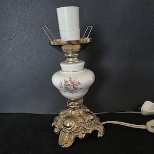 May include: A vintage table lamp with a white ceramic body decorated with pink and purple floral patterns. It has a gold-coloured ornate base and a silver-toned top. The lamp is connected to an electrical outlet.