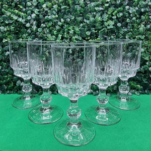 May include: Six clear cut-glass wine goblets. Each glass has a round base, a decorative stem, and a patterned bowl. The glasses are displayed on a green surface with a backdrop of green foliage.