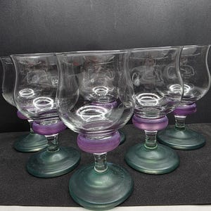 May include: A collection of six clear glass wine glasses. Each glass features a rounded bowl, a purple band on the stem, and a green base. The glasses are displayed in a row, highlighting their design.
