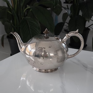 May include: A silver teapot with a rounded body, spout, and handle. The teapot has a lid with a small finial and an ornate handle. An initial is engraved on the side. The teapot is on a white surface.