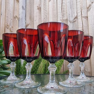 May include: A set of five wine glasses featuring deep red bowls with a vertical arched cut pattern. Each glass has a clear, faceted stem and a circular base, providing a structured look for these drinking vessels arranged on a reflective surface.