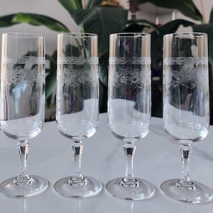 May include: Four clear champagne flutes with etched floral patterns. Each glass has a slender stem and a circular base. The glasses are displayed on a white surface. The flutes are approximately 20 cm tall.