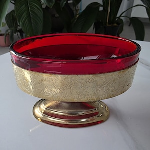 May include: A red glass bowl with a gold-coloured metal base. The bowl has a wide, shallow design. The gold band around the bowl features an embossed floral pattern. The bowl sits on a round, gold-coloured pedestal base.