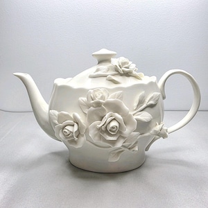 May include: A white ceramic teapot with a lid and handle. The teapot is decorated with raised rose floral designs. The spout and handle are curved. The teapot is a decorative item for serving tea.