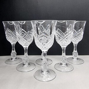 May include: Six clear crystal wine glasses with detailed diamond and floral cut patterns. Each glass has a round base, a decorative stem, and a flared bowl. The glasses are arranged in a row against a dark background.