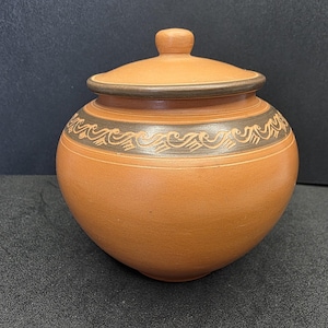 May include: A terracotta-coloured ceramic jar with a lid. The jar has a rounded body and a dark brown band with a decorative wave pattern around the middle. The lid has a small knob on top.