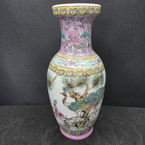 May include: A decorative ceramic vase with a flared top and a wide base. The vase features a hand-painted design of a peacock, flowers, and trees in shades of pink, blue, green, and yellow. The vase is approximately 25 cm tall.