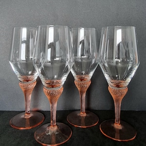 May include: Four clear wine glasses with pink stems and bases. The stems have a textured, twisted design. The glasses have a faceted bowl shape. The glasses are arranged in a row against a dark grey background.
