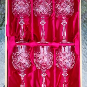 May include: A set of six clear crystal wine glasses with a diamond-cut pattern on the bowls and slender stems, arranged in a rectangular box lined with bright pink satin fabric. Each glass features a small square label with the text HOYA.