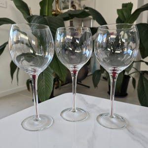 May include: Three clear wine glasses with a red accent on the stem. The glasses have a rounded bowl and a long, slender stem. The glasses are displayed on a white surface, with green plants in the background.