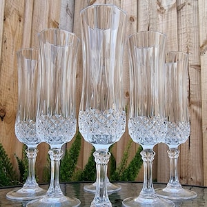 May include: Six clear crystal champagne flutes with a diamond-cut pattern on the lower bowl and a slender stem. The glasses are arranged on a glass surface, with a wooden background visible behind them.