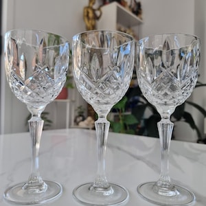 May include: Three clear crystal wine glasses with detailed diamond and leaf-shaped cut patterns. The glasses feature long, elegant stems and round bases. The background is blurred, with hints of greenery and decorative items.