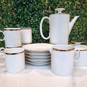 May include: A white porcelain tea set with gold edging. The set includes a teapot, sugar bowl, creamer, cups, and saucers. The cups and creamer have a handle. The set is arranged on a white surface.