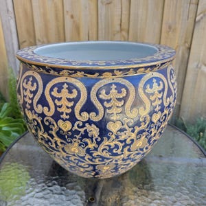 May include: A navy blue ceramic planter with gold ornate detailing. The planter has a wide rim and a repeating pattern of stylized floral and scroll designs. The interior is light blue. The planter is sitting on a glass table.