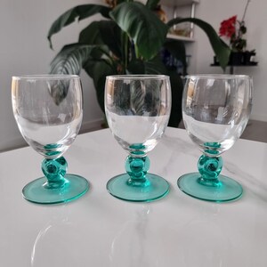 May include: Three clear glass goblets with teal bases and stems. The goblets have a rounded bowl shape and sit on a circular base. The stems feature a decorative, spherical design. The glasses are arranged on a white surface.