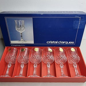 May include: A boxed set of six clear crystal wine glasses, each with a diamond-cut design. The box is blue with the text "Cristal d'Arques" and "6 Longchamp 12 cl."
