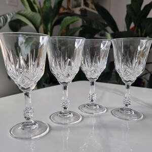 May include: Four clear crystal wine glasses with detailed cut glass patterns. The glasses feature a classic design with a round base, a decorative stem, and a flared bowl. The glasses are arranged on a white surface.