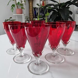 May include: A set of six red wine glasses. Each glass has a clear, circular base, a twisted stem, and a vibrant red bowl. The glasses are arranged on a white surface, highlighting their elegant design and colour.