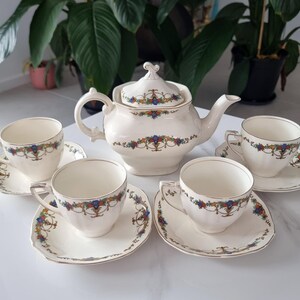 May include: A cream-coloured vintage tea set comprising a teapot with lid, and four teacups with saucers. The porcelain set is decorated with a floral garland design in blue, red, and yellow. The cups and saucers have gold trim.