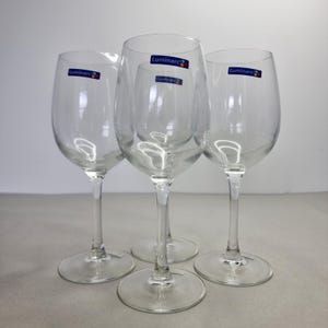 May include: Four clear Luminarc wine glasses with long stems and round bases. Each glass has a blue and white Luminarc sticker. The glasses are arranged on a light grey surface against a white background.