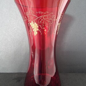 May include: A ruby red glass vase with a gold rim and gold grape vine and leaf design. The vase has a flared top and a slightly curved body. The vase is approximately 25 cm tall.