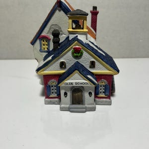 May include: A miniature ceramic schoolhouse with a red and white exterior, blue roof, and a small bell tower. The words "OLDE SCHOOL" are printed above the front door. A small wreath decorates the roof. The building has a chimney and snow-covered roof.