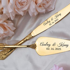 May include: A gold-colored wedding cake knife and server set. The knife and server are engraved with the names "Audrey & Kenny" and the date "02.16.2025". The set has ornate, decorative handles.