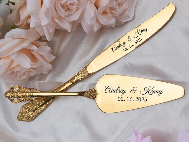 May include: A gold-colored wedding cake knife and server set. The knife and server are engraved with the names "Audrey & Kenny" and the date "02.16.2025". The set has ornate, decorative handles.