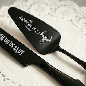 May include: A matte black cake knife and server set featuring engraved white text. The knife reads TILL DEATH DO US PART. The server features a bat graphic, the text The ANDERSONS, and the date 07.05.2026 in a stylized, dripping font.