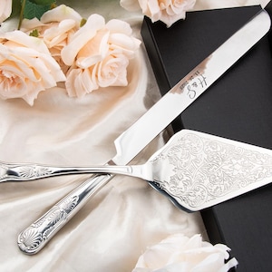 May include: A silver cake knife and server set with ornate floral detailing. The knife is engraved with "H & S" and the date "11. 26. 2023". The set is displayed on a cream-colored fabric with peach roses and a black box in the background.