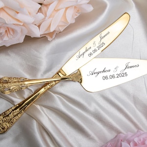 May include: A gold-colored cake knife and server set with ornate handles, engraved with the names "Angelica & James" and the date "06.06.2025". The set rests on a white satin surface, with soft pink and peach roses in the background.