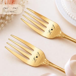May include: Two gold-toned metal forks featuring an engraved design on the handle neck that includes the letters K and J separated by a small heart symbol. The forks have a polished finish and a decorative pattern at the base of the tines.