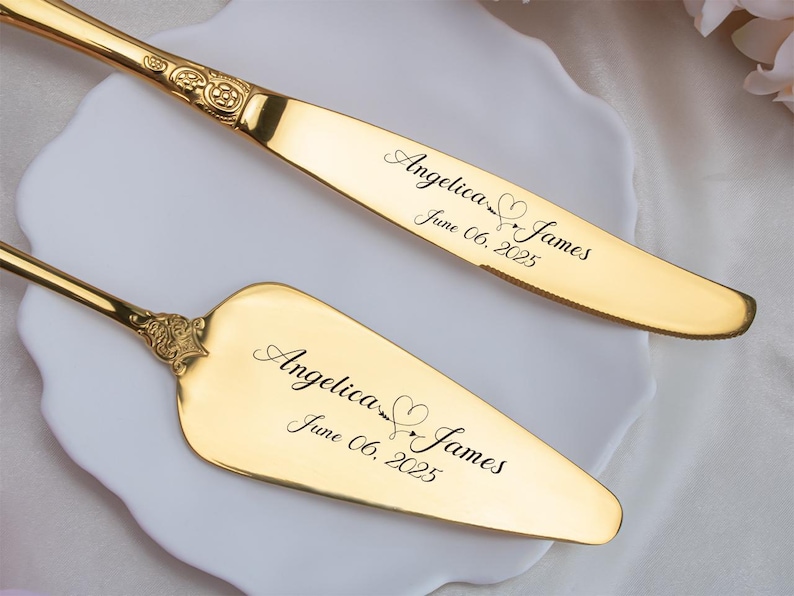 May include: A gold-colored cake knife and server set. The knife and server have ornate handles and are engraved with the names "Angelica & James" and the date "June 06, 2025". The set is displayed on a white plate.