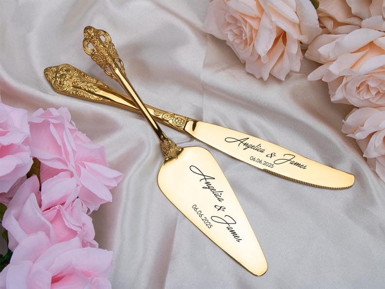 May include: Gold-colored cake server and knife set with ornate handles. The set is engraved with the names "Angelica & James" and the date "06.06.2025". The set is displayed on a white satin surface with pink roses.