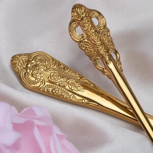 May include: Close-up of two ornate gold-colored serving spoons. The spoons have detailed floral and scrollwork patterns. The spoons are resting on a white satin surface with a blurred pink flower in the foreground. The spoons are likely for serving food.