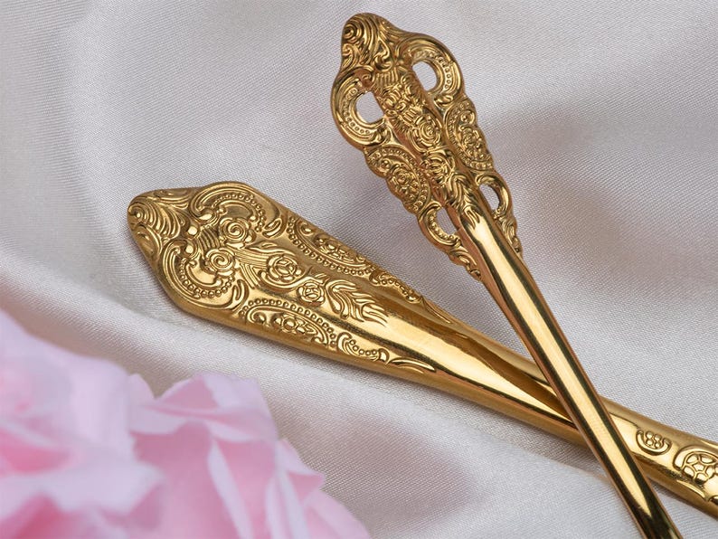 May include: Close-up of two ornate gold-colored serving spoons. The spoons have detailed floral and scrollwork patterns. The spoons are resting on a white satin surface with a blurred pink flower in the foreground. The spoons are likely for serving food.