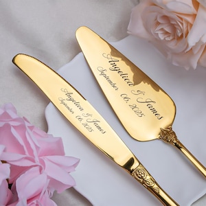 May include: A gold-colored cake knife and server set. The knife and server are engraved with the names "Angelica & James" and the date "September 06, 2025". The set is displayed on a white plate with pink roses in the background.