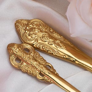 May include: Close-up of two ornate gold-colored serving spoons. The spoons have detailed floral and scrollwork designs. The background is a soft, white fabric with a pink rose in the corner. The spoons are likely for serving food.