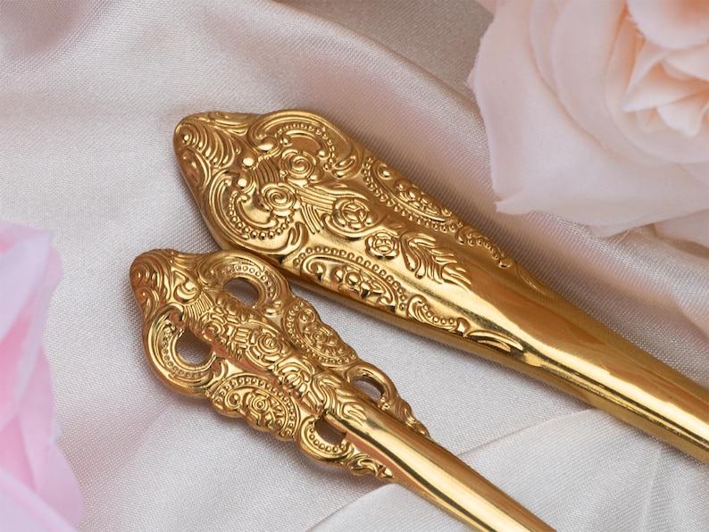 May include: Close-up of two ornate gold-colored serving spoons. The spoons have detailed floral and scrollwork designs. The background is a soft, white fabric with a pink rose in the corner. The spoons are likely for serving food.