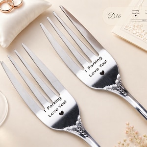 May include: Two silver-toned metal forks featuring an ornate handle design and the engraved text "I Forking Love You!" followed by a small heart symbol. The forks are polished with a reflective finish and have four tines each, showcasing a decorative pattern.