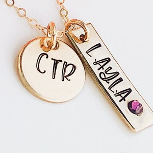 May include: Gold necklace with two charms. One charm is a round disc with the initials "CTR" engraved on it. The other charm is a rectangular bar with the name "LAYLA" engraved on it. The bar charm has a small purple gemstone.